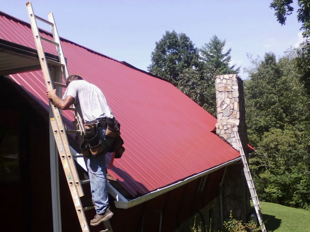 Roofer on a ladder beside a red metal roof and stone chimney — Soffit & Fascia Repair by Roofing Brothers in Ashland City